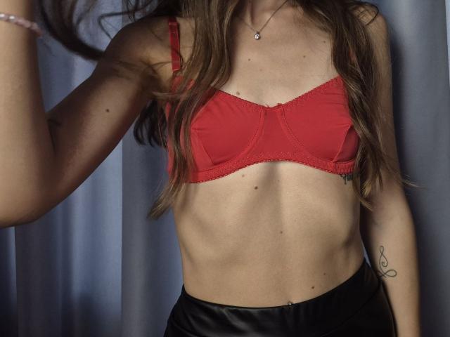 TenerliRize with red bra, showing small tits and slim, toned abs. Long brown hair, black leather pants, tattoo on right arm, and silver necklace. Background is blurred.