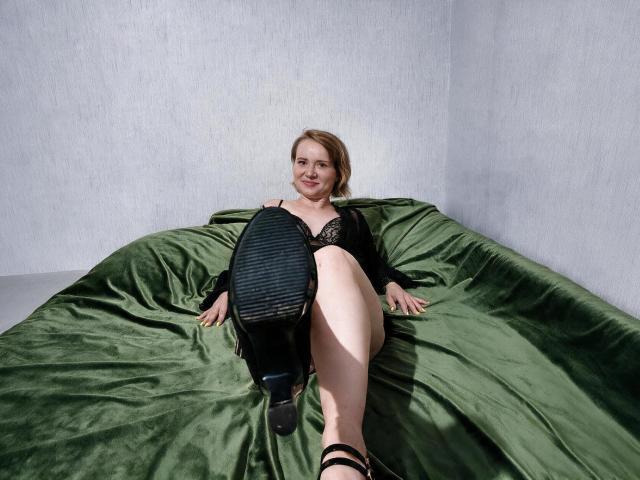 KaterinCold with pale skin, short blonde hair, black lace bra, green bedspread, holding black shoe, smiling, sitting on green bed, wearing black strappy heels.