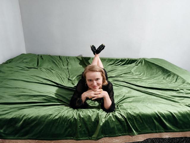 KaterinCold lies on a green bed, her legs up, showing black heels. She's wearing a black shirt, smiling, with her hands under her chin. Her tits are small.
