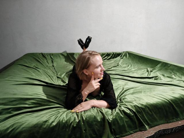 KaterinCold lying on green satin bed, black dress, yellow nails, blonde hair, black high heels in air, hand on chin, looking to the side, gray wall background.