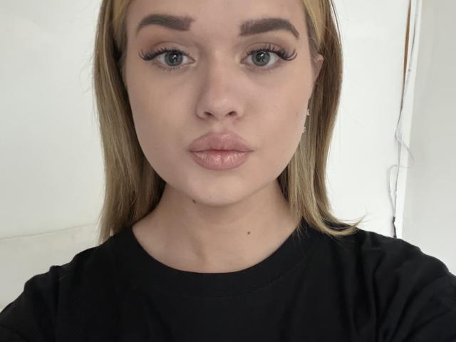 NataliaGschwend with pouty pink lips, blue eyes, and long blonde hair, wearing a black shirt, looking straight at the camera. Her tits are not visible.