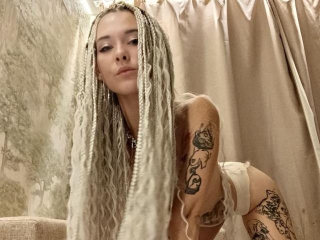 RachelNervis with long white braids, tattoos on arm and thigh, leaning forward in lingerie, showing her ass, in a softly lit, beige room.