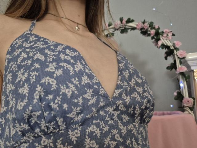 Seliina's tits are prominent in a blue, floral-patterned dress with a deep V-neck, showing a modest amount of cleavage. She's wearing a necklace with a small star.