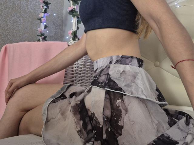 Seliina's ass in a black-and-gray, watercolor-patterned skirt, wearing a black crop top, sitting with her legs spread on a white, tufted chair. Visible red bracelet.
