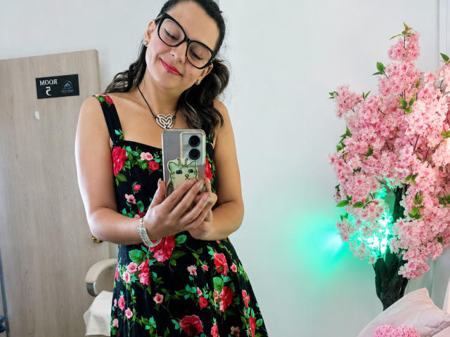 Ariana: Blond, mit Brille, rotem Lippenstift, schwarzem Blumenkleid, hält ein Telefon, trägt eine Halskette und ein Armband, lächelt. Im Hintergrund: rosa Kirschblüten, grünes Licht, eine Holztür und ein Stuhl..