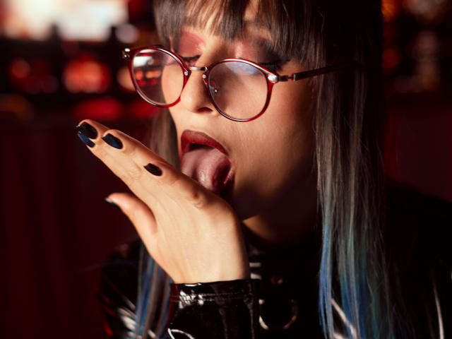 TatsumiLee with dark hair and red glasses, tongue out, black nails, licking her finger, wearing shiny black latex. Red light in background. Slightly erotic.