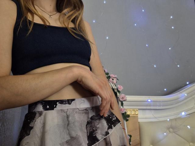 Seliina with blonde hair, wearing a black ribbed crop top, exposing her midriff, and a black and white floral skirt, gently pulling down her skirt.