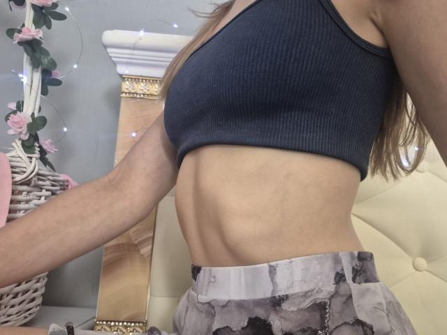 Seliina's slim, toned midsection with visible ribcage, wearing a dark ribbed sports bra and gray, patterned skirt, showing her flat, small ass. Her arm is slightly raised.