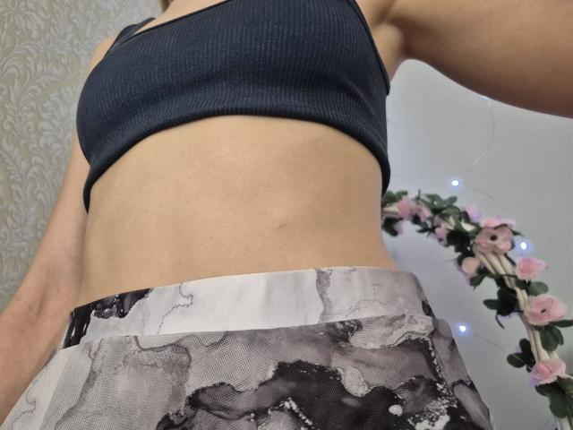 Seliina's midriff, wearing a black sports bra, showing a slim, toned belly, with a gray and black lace skirt, her ass barely visible, a floral garland in the background.