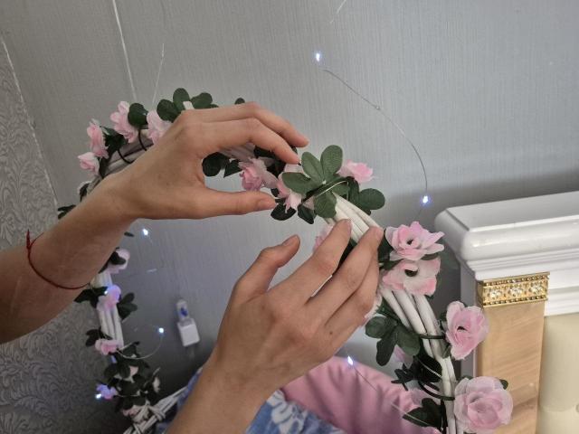 Seliina's hands are decorating a wreath with pink flowers and green leaves, placing a white ribbon. Her red bracelet is on her right wrist.