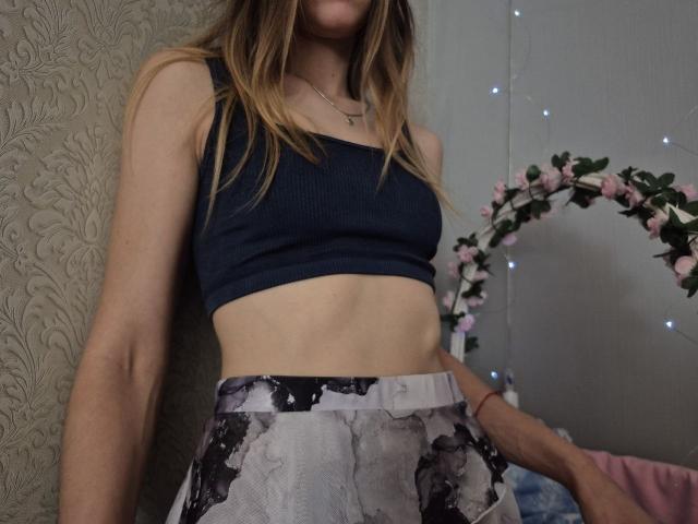 Seliina with slim arms, wearing a navy blue sports bra and floral-patterned skirt, showing her flat abs and small, perky tits. Blonde hair, neutral skin, in a dimly lit room.