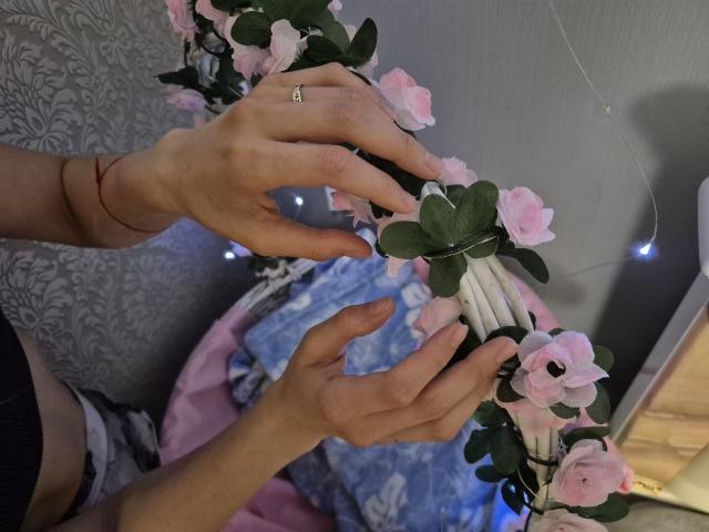Seliina's hands hold a pink rose bouquet with green leaves, showing a ring on her left hand, in a dimly lit room with string lights.