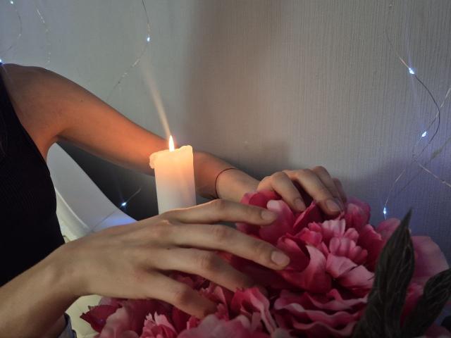 Seliina's hands gently holding pink flower petals, candle flame flickering, soft light, her wrist with red string bracelet, subtle glow from string lights in background.