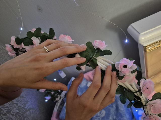 Seliina's hands arranging pink rose and green leaf bouquet, wearing a silver ring, against a white wall with string lights, on a blue-patterned surface.