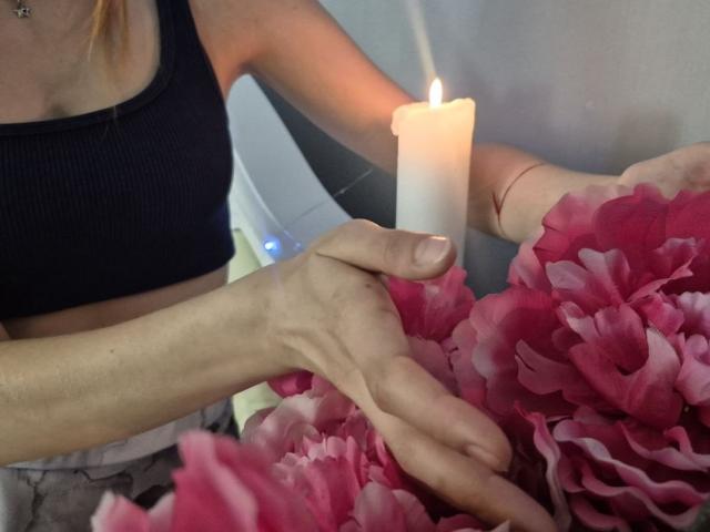 Seliina's hands holding pink flowers next to a burning white candle, wearing a black sports bra, slim arms, light skin, visible wrist tattoo, subtle necklace.