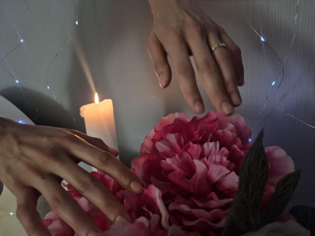 Seliina's hands with a ring on her left ring finger, gently touching pink peony flowers and a lit candle, string lights in the background.