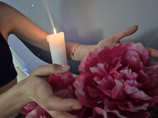 Seliina's arm holds a white candle with a lit flame, while her other hand touches pink peony flowers. Seliina's wrist has a red string bracelet.