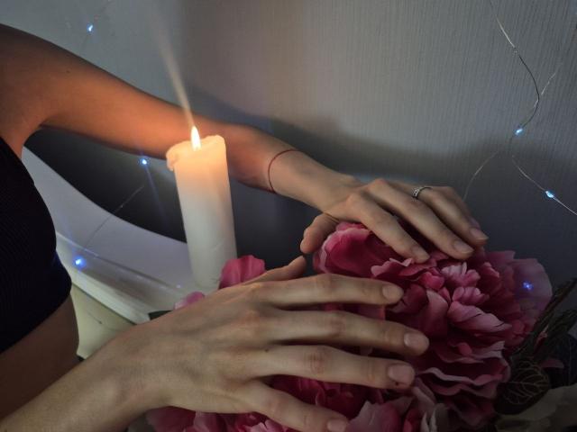 Seliina's hands on pink flowers, next to a lit candle, wearing a black top, ring on right hand, thin red bracelet, subtle fairy lights in the background.