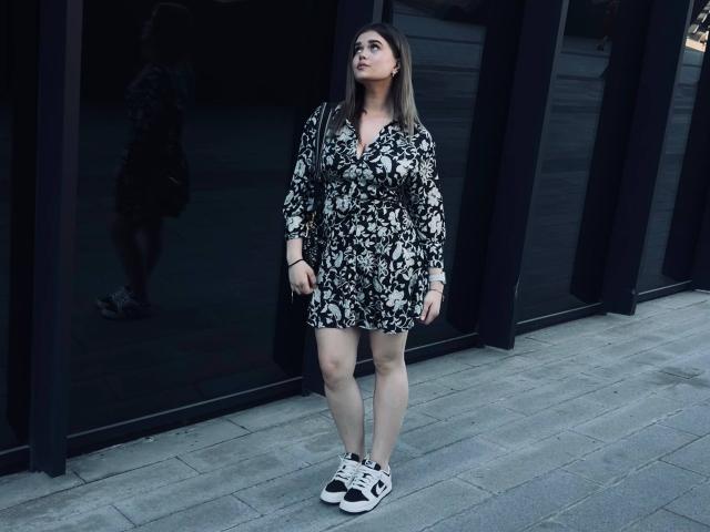 NataliaGschwend with long brown hair, wearing a black and white floral dress, standing in front of a dark glass building, looking up, in white and black sneakers.