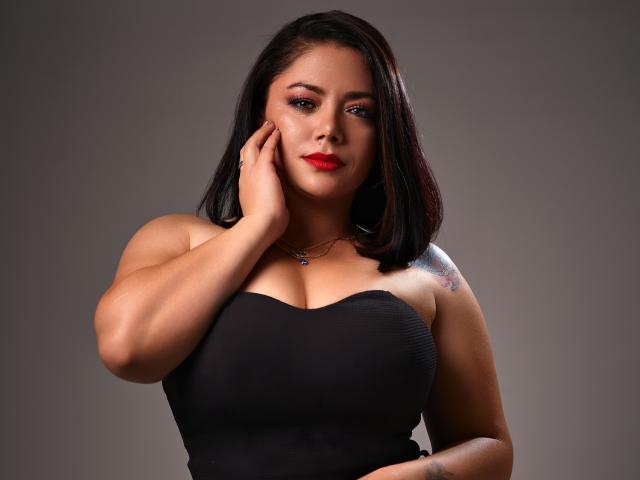 SamanthaVenus with black shoulderless dress, red lipstick, black hair, hand on cheek, tattoo on right shoulder, medium tits, brown skin, gray background, confident expression.