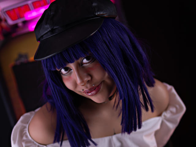 CarlyBlaze, avec ses cheveux bleus, son chapeau noir et son piercing au nez, souriant avec des lèvres brillantes, portant un haut blanc à une épaule qui dévoile sa poitrine, et un collier noir..