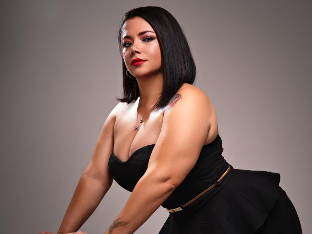 SamanthaVenus a un corps voluptueux et sexy, avec de grandes seins, et elle porte une robe noire sans bretelles, une coupe de cheveux courte noire, du rouge à lèvres rouge, et un tatouage sur son épaule droite..
