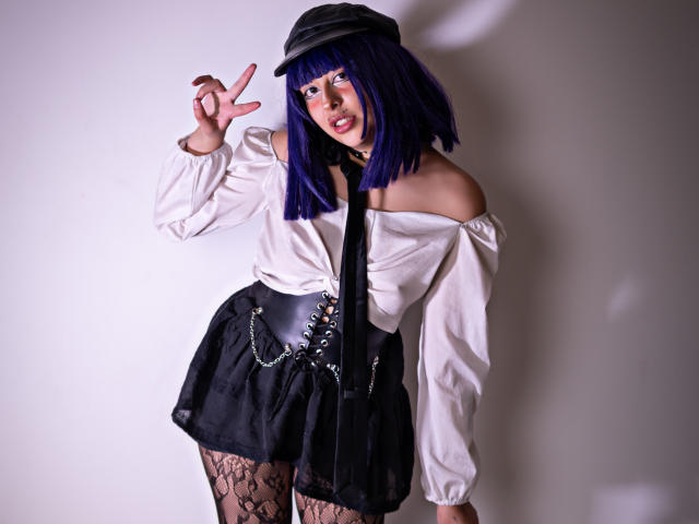 CarlyBlaze, avec une coupe de cheveux courte bleue, portant une casquette noire, une chemise blanche à épaules dénudées, une jupe noire avec des chaînes, des bas en dentelle, et faisant un geste de la main typique du rock-and-roll. Elle a un joli visage..