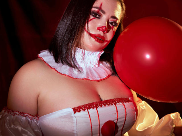 SamanthaVenus met rode lippenstift, bloedige make-up, een wit en rood kostuum, grote borsten, die een rode ballon vasthoudt, donkerrode achtergrond, kort zwart haar, witte gerimpelde kraag, een rood hart op de borst..