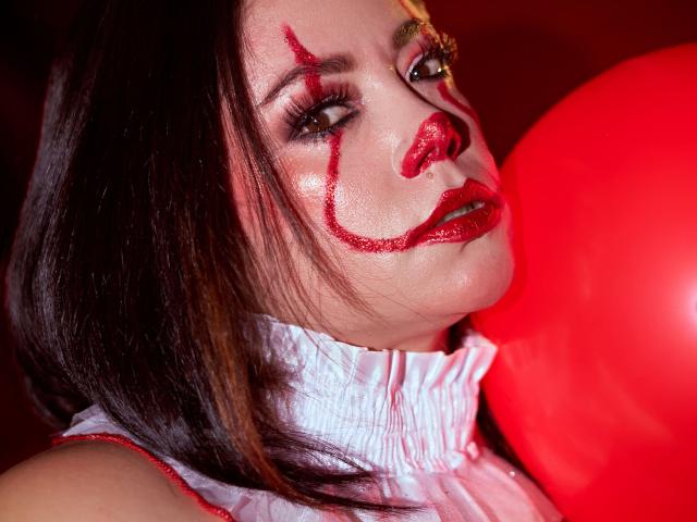 SamanthaVenus met rode make-upstrepen op haar gezicht, glanzende rode lippen, een wit gerimpeld shirt, een rode ballon en donkerbruin haar, die lichtjes omhoog kijkt. Haar borsten zijn gedeeltelijk zichtbaar..