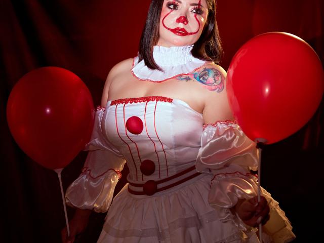 SamanthaVenus, una ragazza formosa con un seno prosperoso, indossa un vestito da clown bianco, tiene in mano palloncini rossi, ha un tatuaggio sulla spalla destra ed ha il trucco da clown rosso..