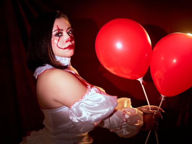 SamanthaVenus mit rotem Make-up, einem weißen, gerafften Kleid und weißen Handschuhen, die zwei rote Luftballons hält und mit einem verführerischen Ausdruck zurückblickt. Ihr Hintern ist leicht sichtbar..
