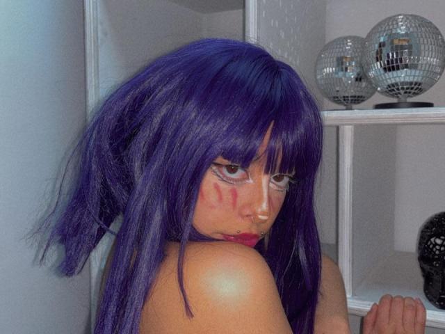 CarlyBlaze, com cabelo roxo, usando maquiagem clara nos olhos e tinta vermelha no rosto, mostrando os ombros, olhando por cima do ombro, parada perto de bolas de espelhos, segurando uma prateleira..