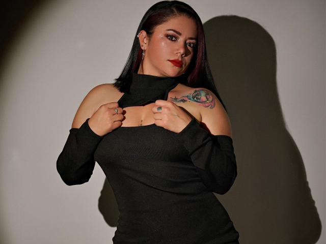 SamanthaVenus met rode lippenstift, een zwarte top met open schouder, die de kraag vasthoudt, zwarte armen, een tatoeage op de rechter schouder, een schaduw op de muur, donker haar, een zelfverzekerde uitdrukking, toont haar borsten..