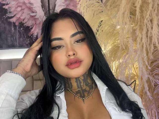 LairaHearvy has long black hair, thick eyebrows, and a tattooed neck with a chain. She's wearing a white shirt, looking at the camera with pink lips.