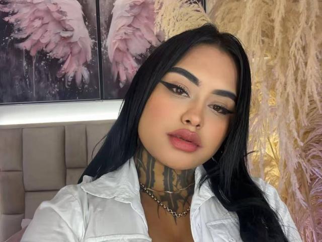 LairaHearvy has dark hair, full pink lips, thick black eyebrows, and a tattooed neck. She's wearing a white shirt, and has a chain necklace. Her boobs are partially visible.