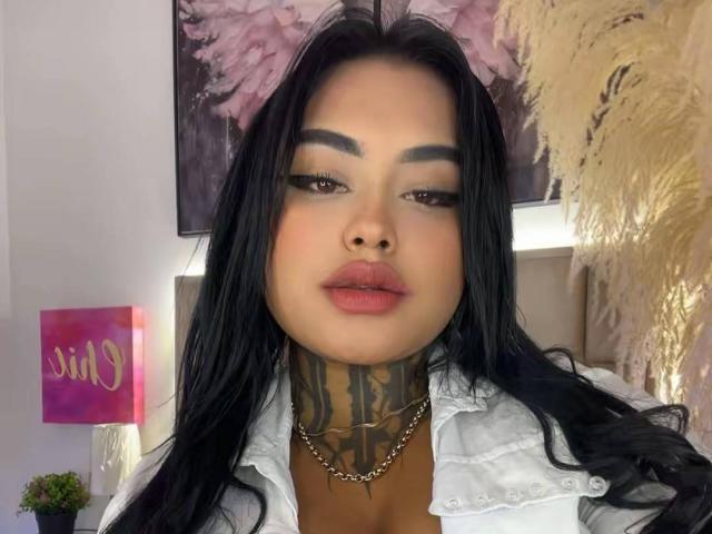 LairaHearvy has long black hair, thick eyebrows, and a tattooed neck, wearing a white shirt with her plump lips slightly parted, and a chain necklace.