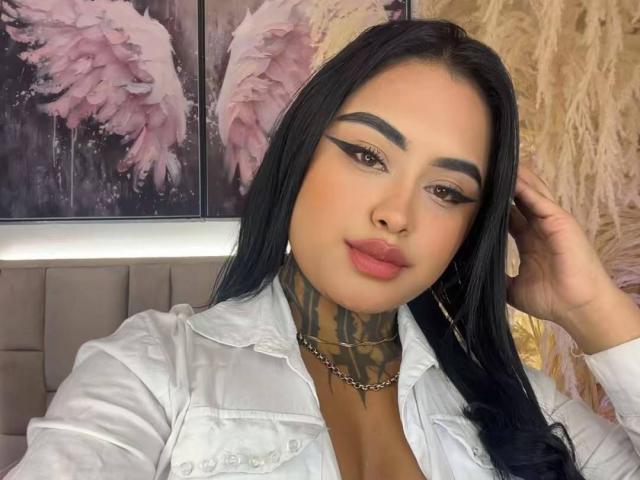 LairaHearvy with black hair, thick eyebrows, winged eyeliner, pink lipstick, white shirt, tattooed neck, hand in hair, and hoop earrings. Boobs visible under shirt.