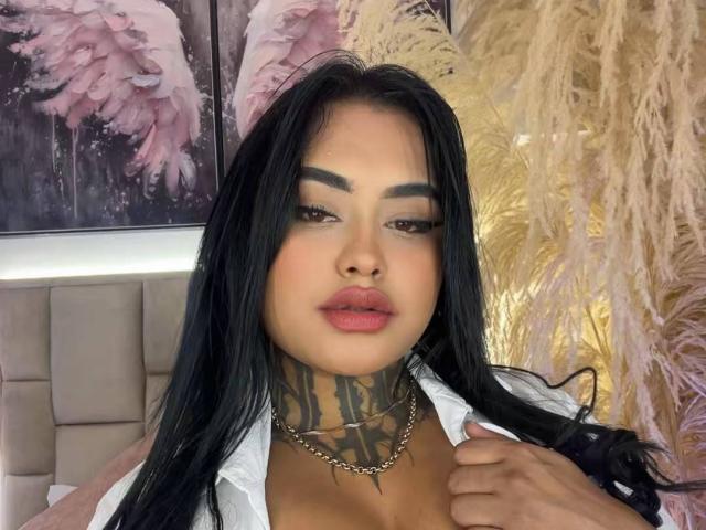 LairaHearvy has long black hair, tattooed neck, full pink lips, and a silver necklace, wearing a white shirt, looking down with a serious expression. Her tits are partially visible.