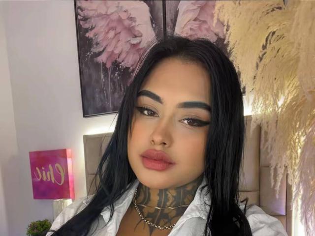 LairaHearvy with black hair, full lips, tattoos on her neck, and bold eyebrows, wearing a white shirt, and a silver chain, looking at the camera.