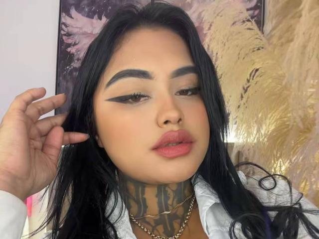 LairaHearvy has black hair, thick eyebrows, bold eyeliner, pink lips, tattoos on her neck, and wears a white shirt, with a hand touching her hair.