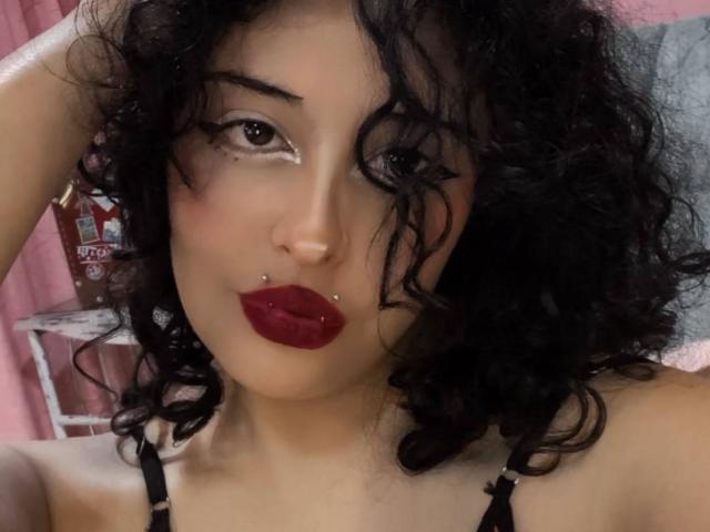 CarlyBlaze with curly black hair, red lipstick, and a nose piercing, wearing a black strap, has white eye makeup and a lip piercing, looking at the camera.