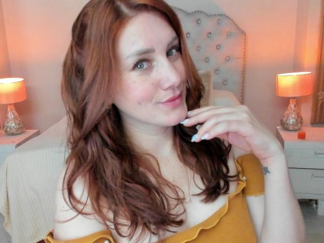 KatyaRogersX with wavy red hair, off-shoulder yellow top, pale skin, and a slight smile, touching her chin with a hand, showing her tits and a small tattoo.