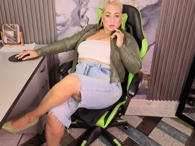 GianellaCox sits in a green and black chair, wearing a green jacket, white tube top, and ripped denim skirt, with her hand near her pink-lipped, slightly open mouth.