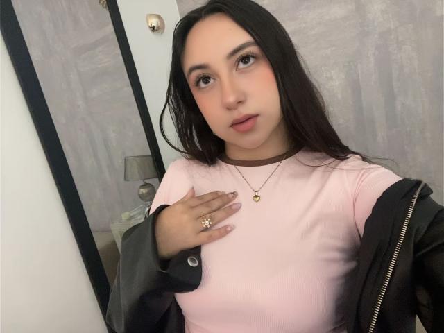 RosalieDupont with long black hair, pink top, black jacket, gold necklace, and ring, hand on chest, looking at camera, soft pink lipstick, light skin, medium tits.