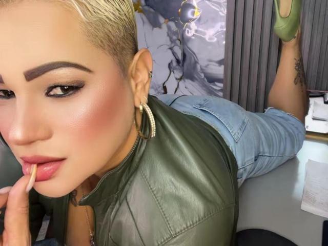 GianellaCox with short blonde hair, green leather top, blue denim shorts, green high heels, finger in mouth, bold eyebrows, pink blush, hoop earrings, tattoo on right ankle.