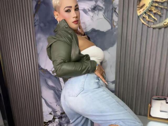 GianellaCox has blonde hair, green jacket, white tube top showing big tits, and tight blue jeans highlighting her ass. She's standing with a sultry look, hands on her hip.
