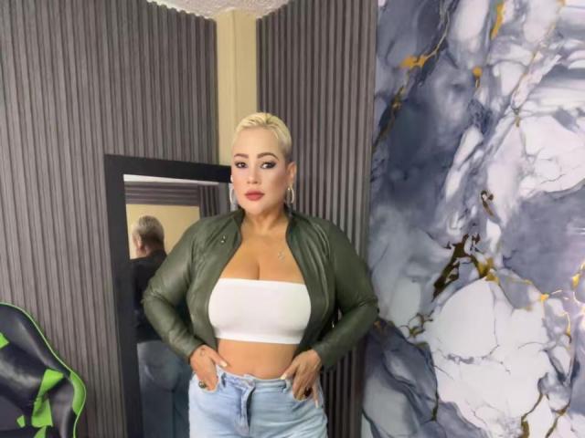 GianellaCox with blonde hair, green jacket, white tube top showing big tits, and blue jeans, standing in front of a mirror, looking at herself with hands on hips.