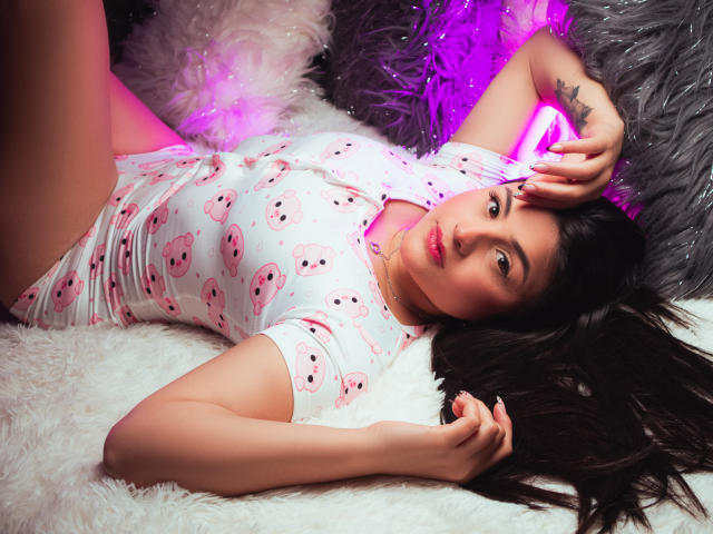 SaraWestt lying on white fur, wearing pink skull-patterned bodysuit, with pink neon light, arm tattoo, long brown hair, and hand on head, showing her ass.