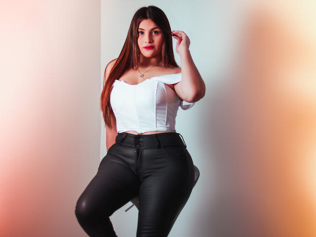SaraWestt with long brown hair, white off-shoulder top, and black leather pants, standing, right hand in hair, red lipstick, small necklace, white wall, soft lighting, sexy.