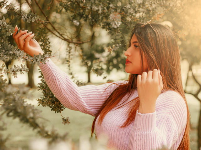 SaraWestt, avec ses longs cheveux bruns, portant un pull rose en tricot, touche une branche d'arbre avec sa main droite, la lumière du soleil filtrant à travers les feuilles, une douce lueur sur son visage..